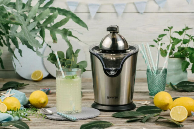 Gorenje cediljka za citruse na drvenm stolu, okružena limunom i čašom sa limunadom, u zelenilom obogaćenom okruženju.